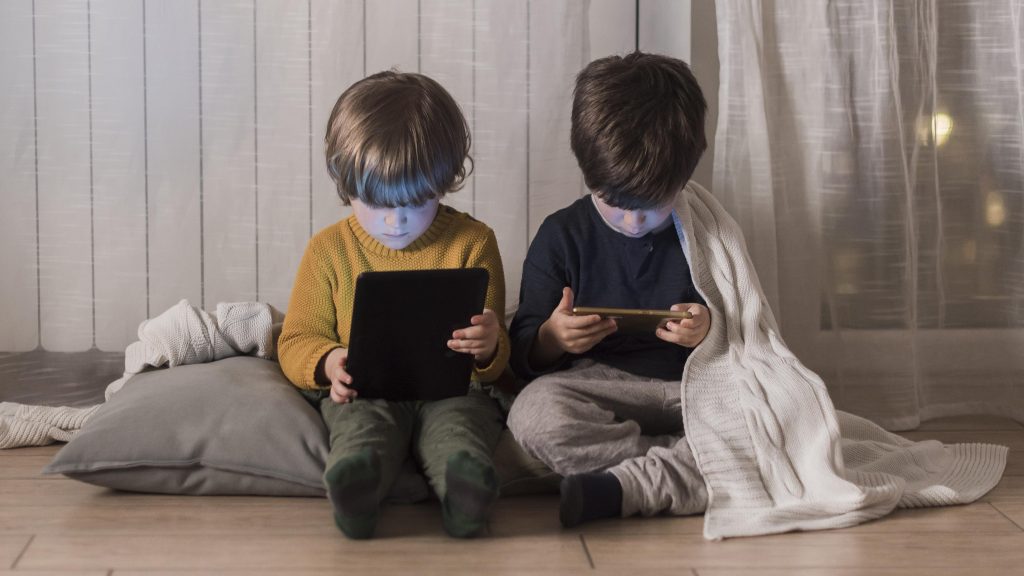 Two young children sitting indoors, absorbed in tablet and smartphone screens, surrounded by soft furnishings.